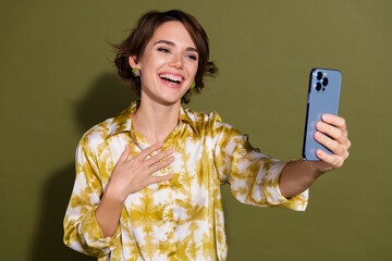 Smiling young woman in elegant blouse takes selfie with smartphone on khaki background showcasing...