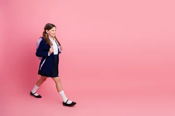 Gordijnen Charming schoolgirl in uniform walking confidently carrying a backpack against a stylish pink background © deagreez