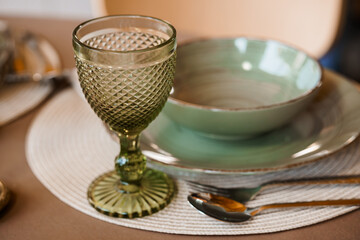 Elegant Table Setting with Green Glassware and Ceramic Plates. Rustic Dining Scene, Brown Tones, Textured Placemats and Golden Cutlery.