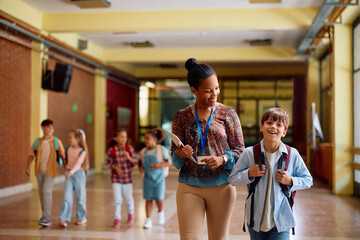 Happy black teacher and elementary student walking through hallway.