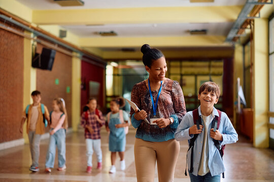 Happy black teacher and elementary student walking through hallway.