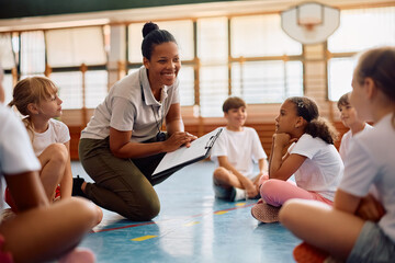 Happy black female coach with group of children during PE class at school gym.
