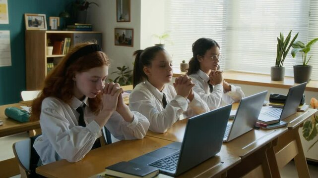 Medium shot of focused diverse student girls in uniforms praying before computer science lesson in modern religious school sitting in front of laptops