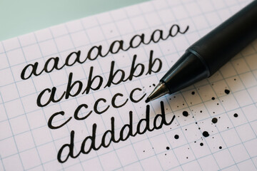Black ink pen writing cursive lowercase letters on grid notebook paper with ink splatters and close up detail of calligraphy practice