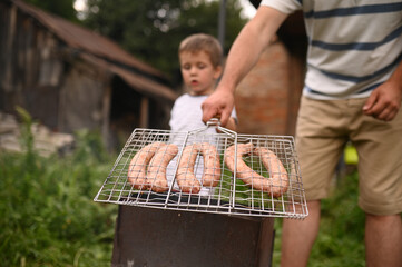Father holding grill grate with sausages close-up hand. Family time, involved parenting, picnic...