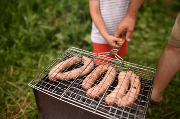 Child holding grill grate with sausages close-up hand. Family time, involved parenting, picnic barbecue outdoor recreation with children
