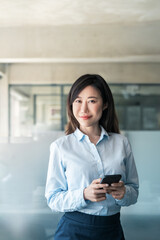 Vertical portrait of asian Korean business woman manager using cell phone mobile trade app standing in office. Middle age female ceo businesswoman working on smartphone cellphone looking at camera
