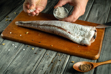 Hands sprinkle salt onto a long fish placed on a cutting board. Various spices are set nearby,...