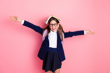 Confident schoolgirl wearing a uniform with a book on head showing happiness against pink background