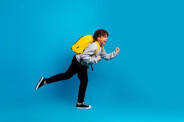 Energetic young schoolboy with a yellow backpack joyfully running against a bright blue background...
