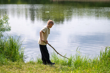 Boy standing on lakeside with a long stick, playing near the water in nature on a summer day