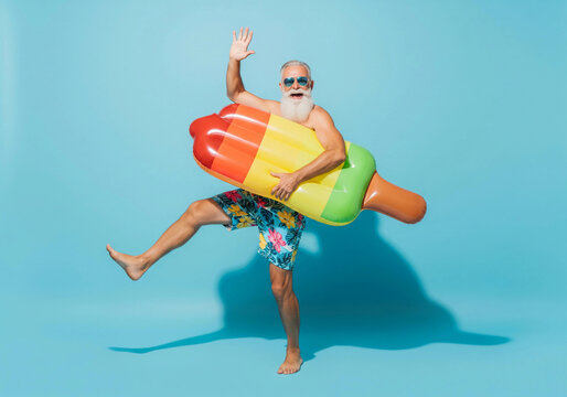 Elderly man with a white beard wearing sunglasses and colorful shorts poses playfully with an inflatable pool float on a blue background, embracing fun summer vibes and tropical vacation energy.

