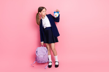 Young schoolgirl holding an alarm clock while posing with a backpack against a pink background