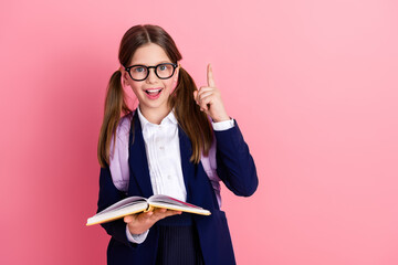 Smiling schoolgirl in formal uniform reading a book and pointing upwards on pink background...
