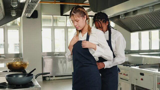 In a professional kitchen at a restaurant a girlfriend chef assists her woman friend tying apron strings before their food education. Two women cheerful and standing preparing for service.
