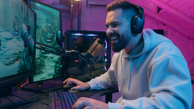 Smiling happy Caucasian addicted male gamer guy man streamer play at computer internet virtual gaming at home web battle online playing video game at dark evening with microphone and headset laughing