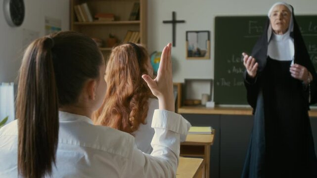 Mature Caucasian nun teacher in veil having lesson in religious school with student girls in white uniforms answering her questions sitting at desks in modern classroom with crucifix on wall