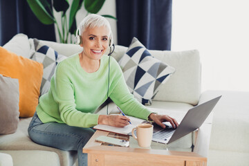 Smiling mature woman wearing a green sweater with headset working from home on a laptop, seated on...
