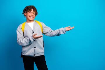 Smiling boy with a backpack gesturing to the side on a blue background, dressed in casual school...
