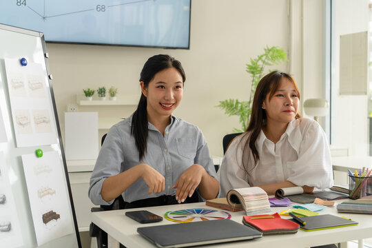 Two Asian women working together in a bright, modern studio.while using a laptop and smartphone. The creative workspace is filled with colorful samples for seasonal palette selection.