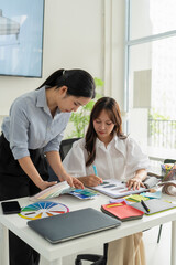 Obraz premium Two Asian women working together in a bright, modern studio.while using a laptop and smartphone. The creative workspace is filled with colorful samples for seasonal palette selection.