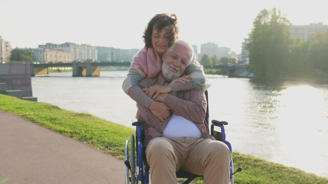 Old man in wheelchair walking with caregiver senior woman on road in park. Elderly family couple woman supporting embracing paralyzed man in chair for people with disability outdoor. Rehabilitation