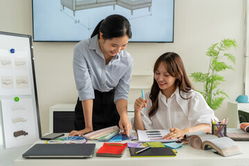 Obraz premium Two Asian women working together in a bright, modern studio.while using a laptop and smartphone. The creative workspace is filled with colorful samples for seasonal palette selection.