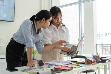 Two Asian women working together in a bright, modern studio.while using a laptop and smartphone....