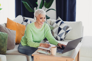 Confident woman with headphones using a laptop while taking notes in a cozy living room setting,...