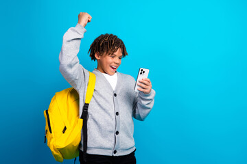 Joyful schoolboy with a backpack celebrating success while using a smartphone on a vibrant blue background
