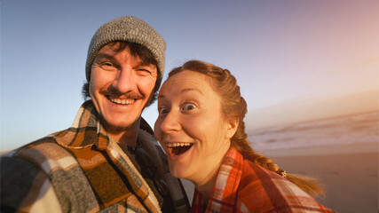funny Couple in warm Plaid Shirts Embracing and Taking Selfie by the Ocean