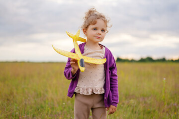 Young girl standing in a field at sunset, gently holding a yellow toy airplane. She wears a purple...