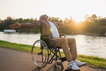 Happy senior man in wheelchair raised hands enjoying freedom walking on road in city park outdoor. Old happy man in chair for people with disability has a hope. Disability disease rehabilitation