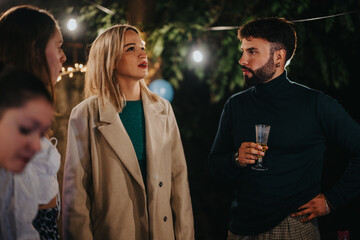 An evening celebration featuring a group of friends conversing, enjoying drinks, and a festive atmosphere outdoors with decorative lighting and casual attire.