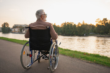 Sad senior man sitting in wheelchair walking on road in city park outdoor, back view. Old paralyzed man in chair for people with disability feel depressed. Lonely elderly sick man sits on wheelchair