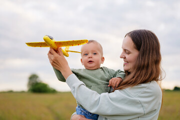 A mother holding her toddler son while they play with a yellow toy airplane in a field, sharing a...