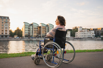 Sad senior woman sitting in wheelchair walking on road in city outdoor, back view. Old paralyzed lady in chair for people with disability feel depressed. Lonely elderly sick woman sits on wheelchair