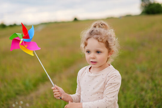 A cute little girl with blonde curly hair holds a colorful pinwheel, looking curiously ahead in a grassy field.