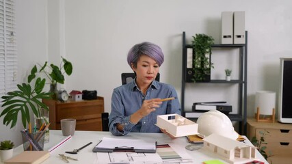 Experienced female architect analyzing paper plans and taking notes beside 3D model on desk indoors - Powered by Adobe