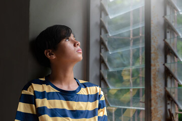 A teenage boy in a striped shirt gazes thoughtfully out of a louvered window, bathed in soft...