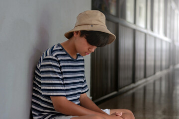 Asian teenage boy wearing a striped shirt and bucket hat sitting alone against a wall, looking down pensively in a quiet hallway.