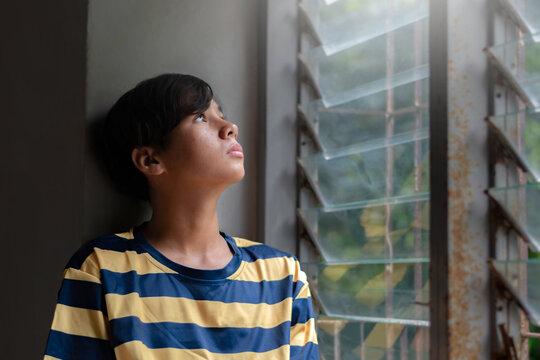 A teenage boy in a striped shirt gazes thoughtfully out of a louvered window, bathed in soft natural light, evoking a mood of solitude and contemplation.