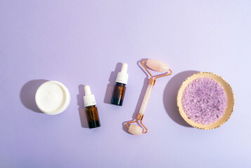 Spa cosmetic set with cream jar, essential oil bottles, face roller and sea salt on lilac background. Top view, copy space