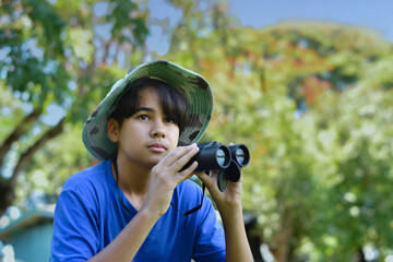 A curious boy in a wide-brim hat looks through binoculars during an outdoor exploration. Surrounded by trees, he enjoys a nature adventure on a sunny day.