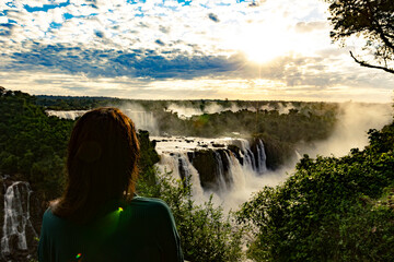 Woman admires the stunning view of Iguazu Falls at sunrise, surrounded by mist and rainforest. The...