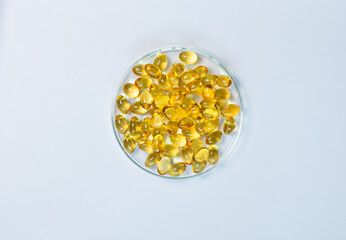 Top-view of yellow and amber supplement capsules in a glass petri dish, isolated on a white background. This shows scientific research, pharmaceutical development for health products like fish oi.