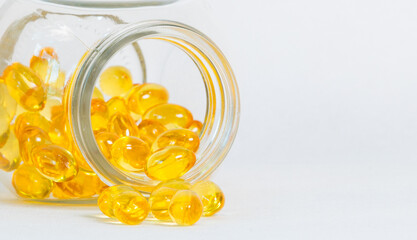 Glass jar with yellow vitamin capsules spilled on white surface. Clean layout with copy space emphasizes softgel fish oil supplements, wellness lifestyle, and modern healthcare concept.