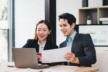 Two Asian business professionals, man and woman, wearing formal suits, collaborate at modern office desk with laptop and financial documents, expressing positive teamwork and focus