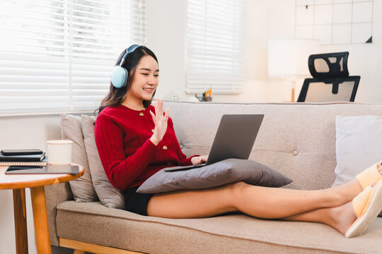 Young woman Asian wearing red sweater sitting on sofa with pillow using laptop wearing headphones smiling and waving during video call relaxed home environment natural light
