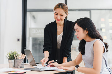 Two Asian women working together in modern office, one standing and pointing at document while other sits and writes, teamwork and collaboration concept